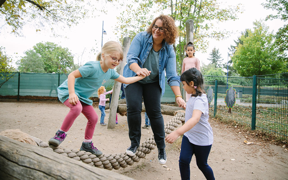 Bild: Eine Erzieherin balanciert auf einem Spielplatz und wird von Kindern geführt.
